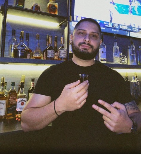 Mike, bartender, behind a lit-up bar holding a shot